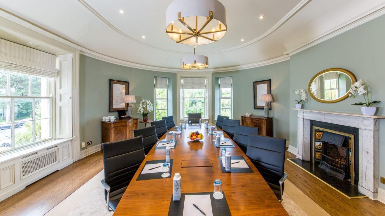 Elegant meeting room with a polished table, plush chairs, fireplace, and large windows overlooking lush greenery, ideal for events.