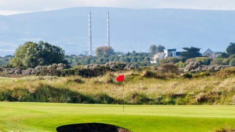 Scenic golf course with rolling green hills, a vibrant red flag, and distant industrial towers under a blue sky.