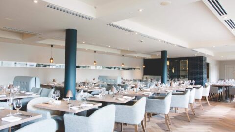 Spacious hotel dining area with elegant grey chairs, polished tables, soft lighting, and large windows for a bright, welcoming experience.