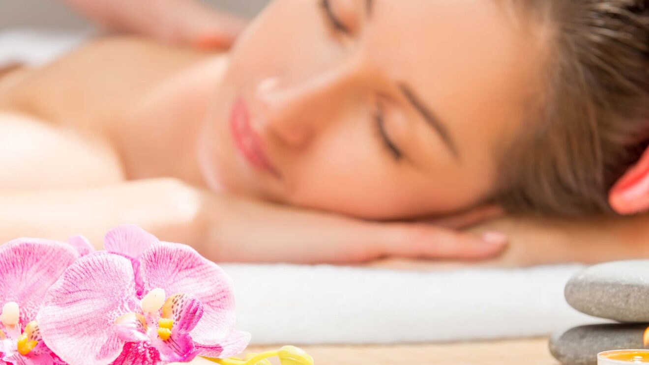 Woman relaxing with massage at luxury hotel spa next to pink orchids and smooth stones, promoting tranquility and wellness.