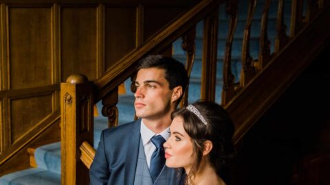 Elegant couple on grand staircase at luxurious hotel, showcasing traditional wood panelling and soft blue carpet.