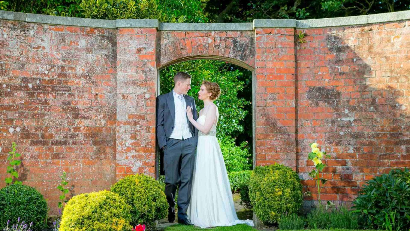 Couple in elegant attire admire lush hotel garden with vibrant flowers and historic brick archway. Ideal wedding location.