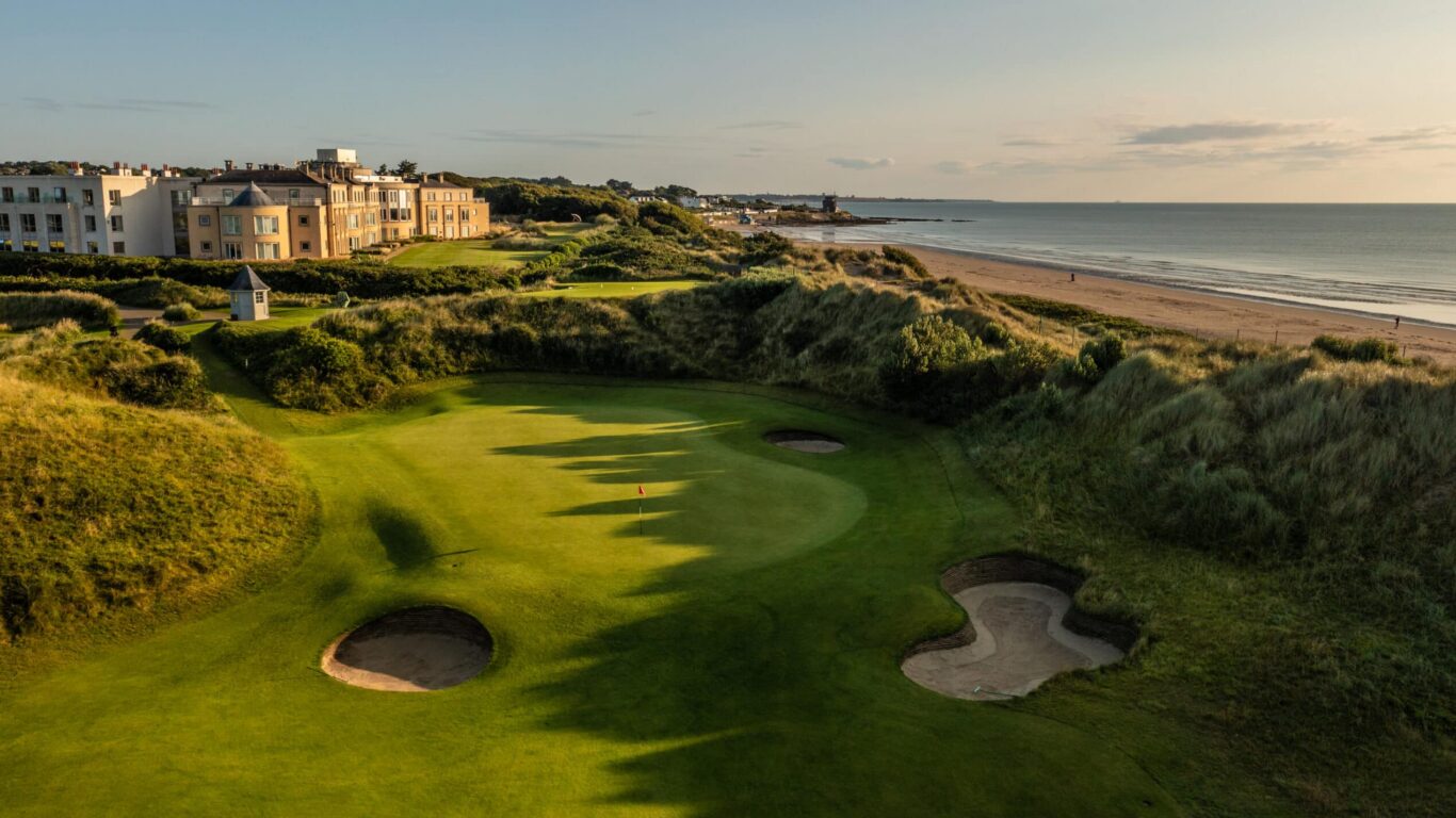 Coastal hotel with lush golf course, surrounded by sandy beaches and greenery, offering serene ocean views at sunset.
