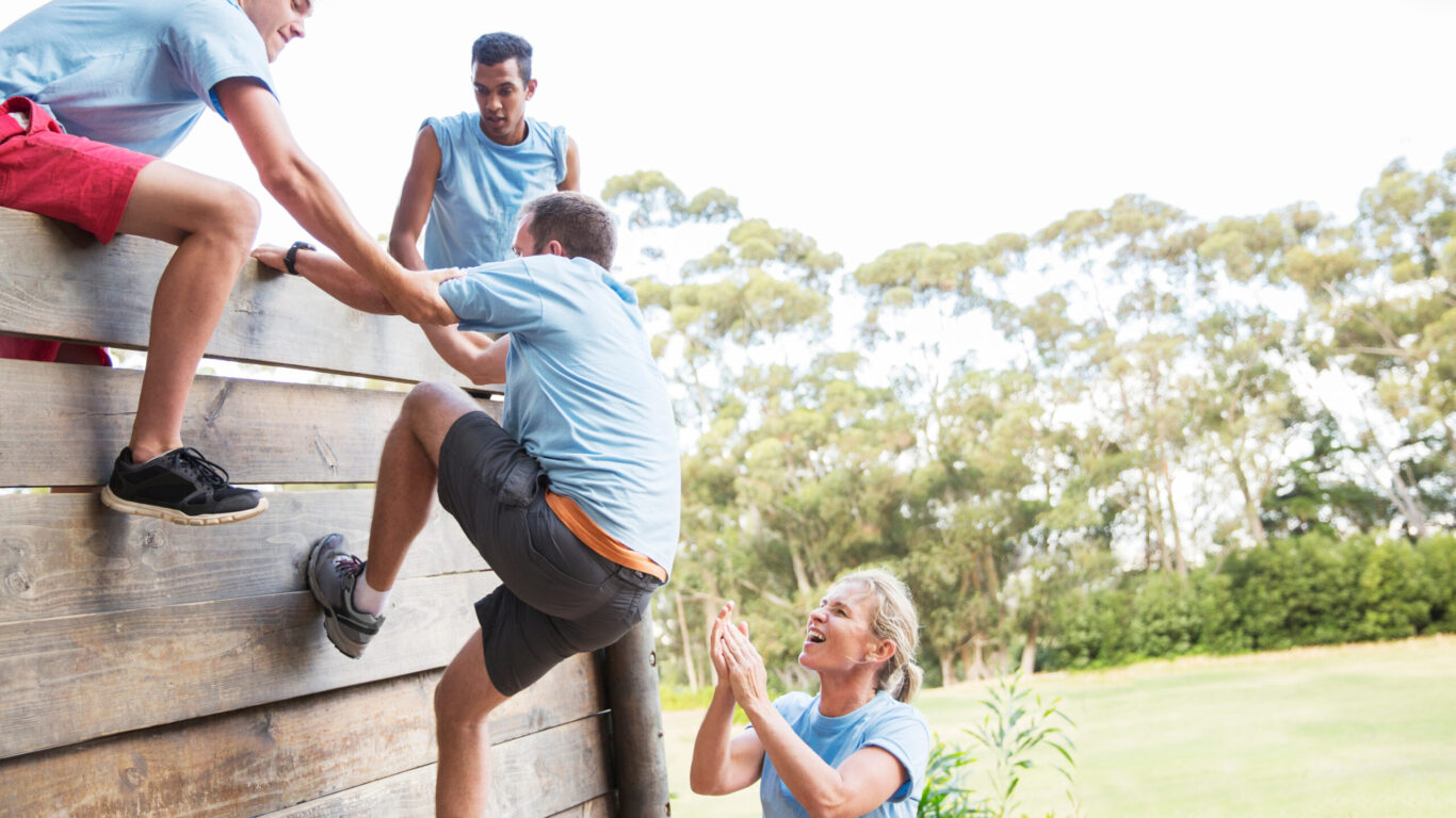 Teammates helping man over wall on boot camp obstacle course