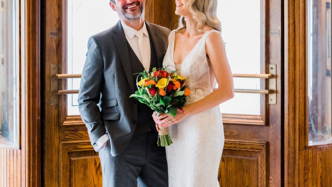 Elegant couple smiling in front of classic wooden hotel entrance, marble floor; perfect for weddings and romantic escapes.