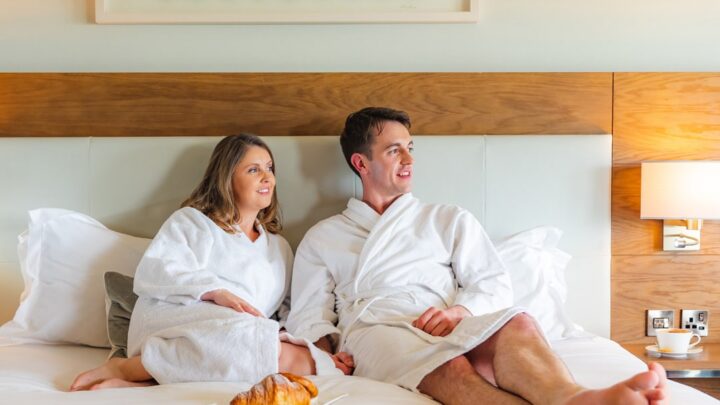 Guests relaxing in plush robes on a king-size bed with a croissant, modern art, and warm lighting creating a cosy atmosphere.