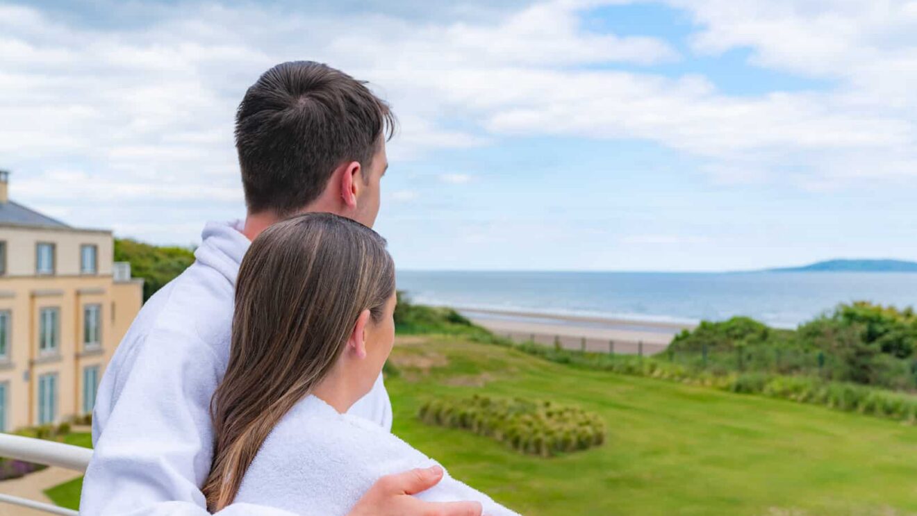 Guests in bathrobes admire ocean view from hotel room balcony, overlooking lush gardens and sandy beach.&nbsp;