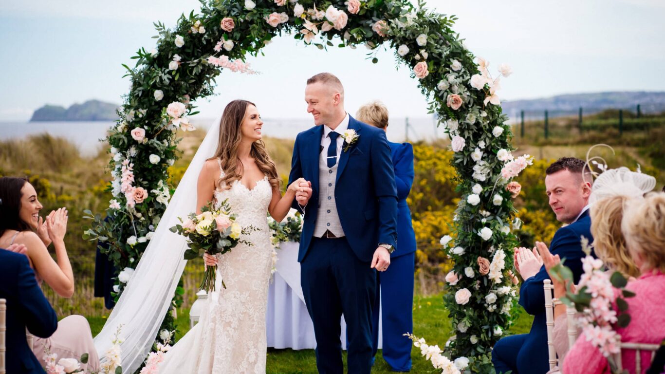 Happy couple under floral arch at seaside garden wedding, with stunning coastal views and lush greenery.