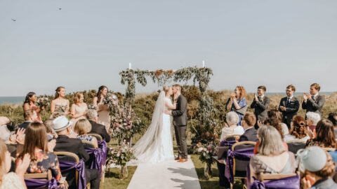 Romantic outdoor wedding ceremony with a joyful couple kissing under a floral arch, guests applauding on a sunny day.