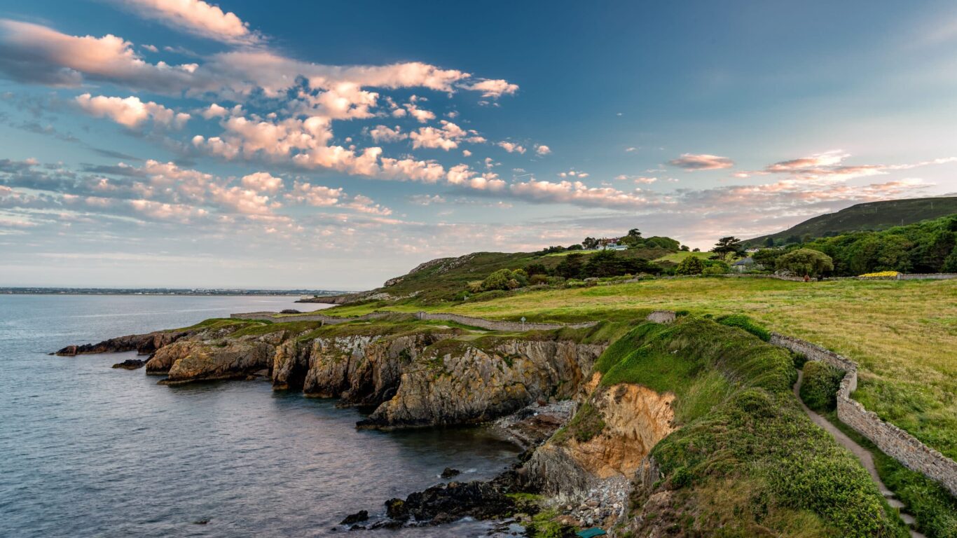 Dramatic coastal cliffs and a scenic walking path in Howth, Dublin, Ireland, with lush green fields, rugged shoreline, and a sky dotted with soft pink clouds during sunset.