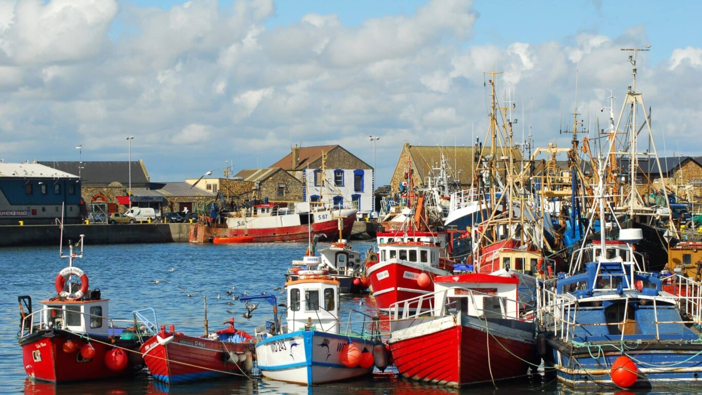 Colourful fishing boats docked at Howth Harbour in Dublin, Ireland, with historic stone buildings and a bright blue sky with scattered clouds in the background.