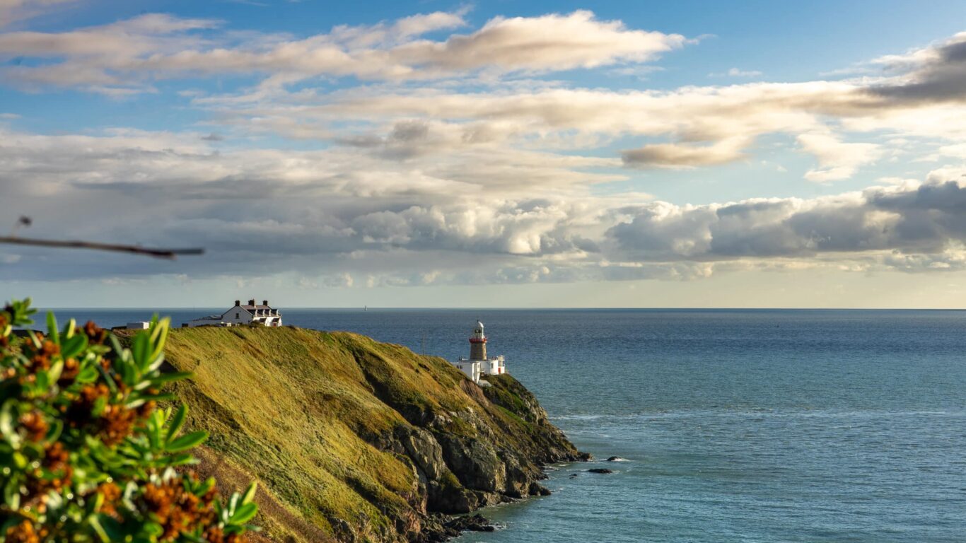 Scenic coastal view of a lighthouse perched on a grassy cliffside overlooking a calm sea under a partly cloudy blue sky, with a white cottage visible further inland and flowering bushes in the foreground.