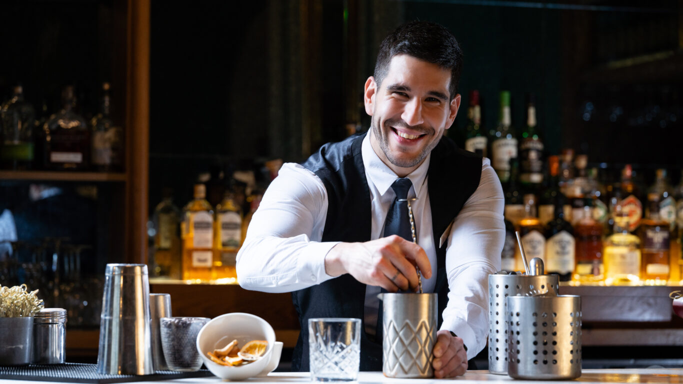 Friendly bartender mixes cocktails at elegant hotel bar with well-stocked shelves, welcoming atmosphere, and craft drinks.