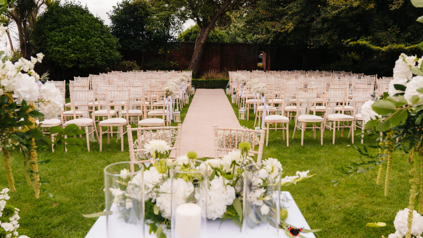 Elegant outdoor wedding setup with white chairs and floral arrangements on lush garden lawn, creating a romantic ceremony space.