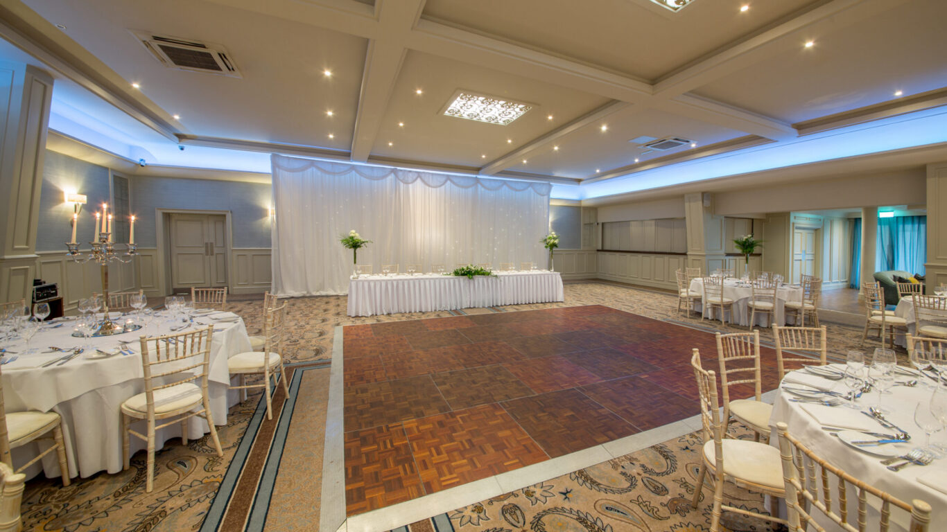 Elegant hotel ballroom with banquet tables set for a wedding, featuring stylish decor, wooden dance floor, and soft lighting.