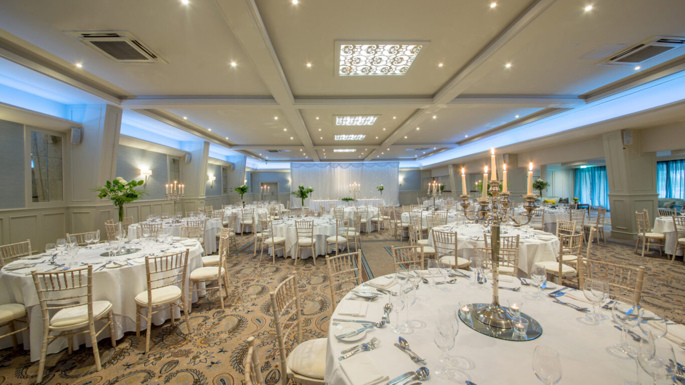 Elegant hotel ballroom set for an event with round tables, white linens, candle centrepieces, and soft ambient lighting.