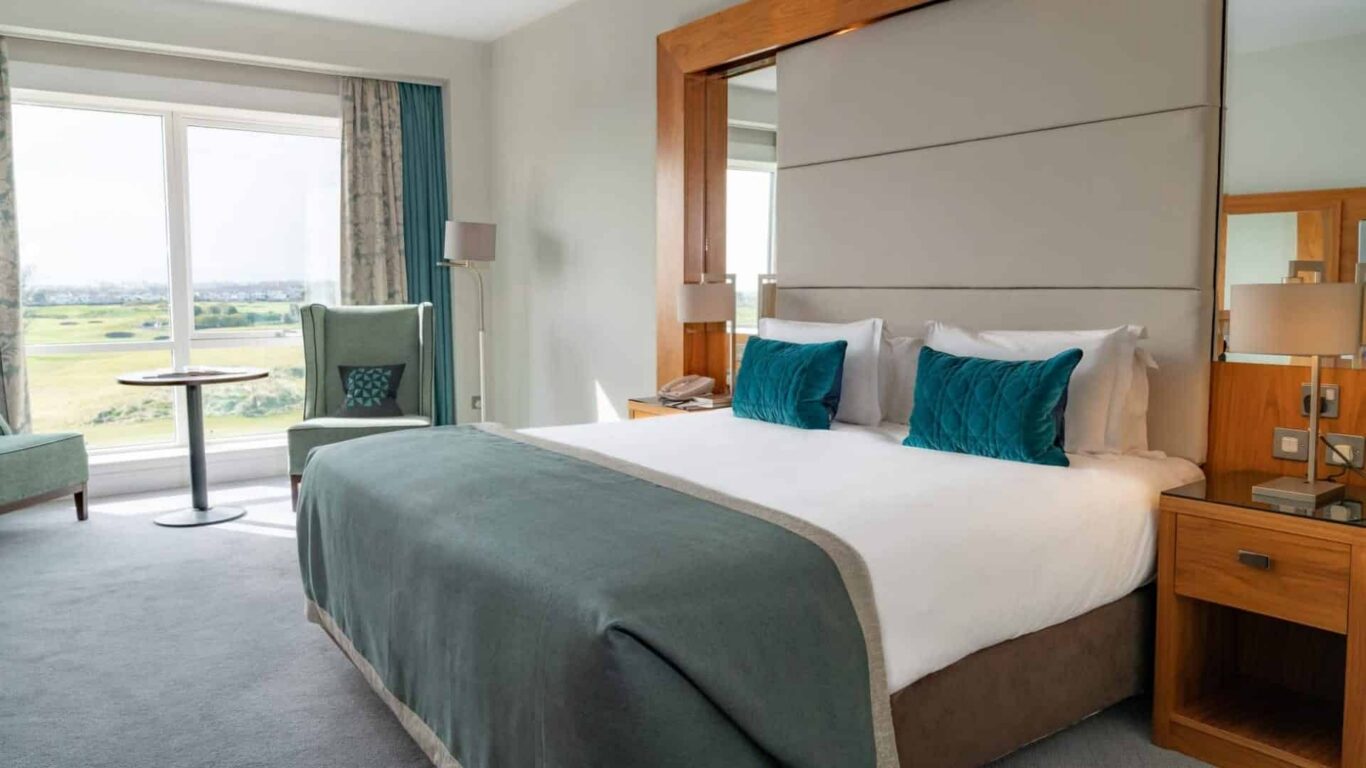 Spacious hotel room with king bed, plush teal pillows, seating area, and scenic countryside view from large window.
