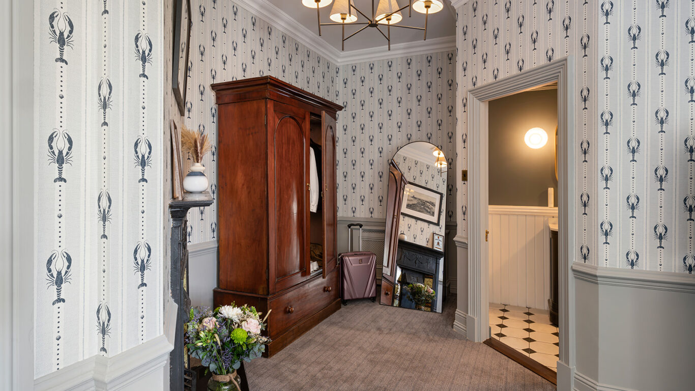 Charming hotel room with elegant wallpaper, wooden wardrobe, mirror, and floral decor, offering a cosy and stylish retreat.