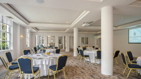 Elegant, spacious hotel conference room with round tables, blue chairs, chic decor, natural lighting, ideal for events.