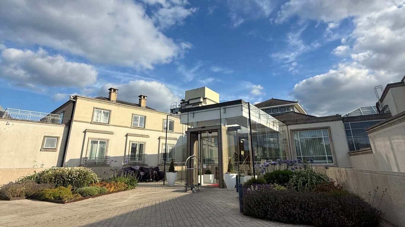 Modern hotel entrance with glass facade, surrounded by lush gardens under a vibrant blue sky.