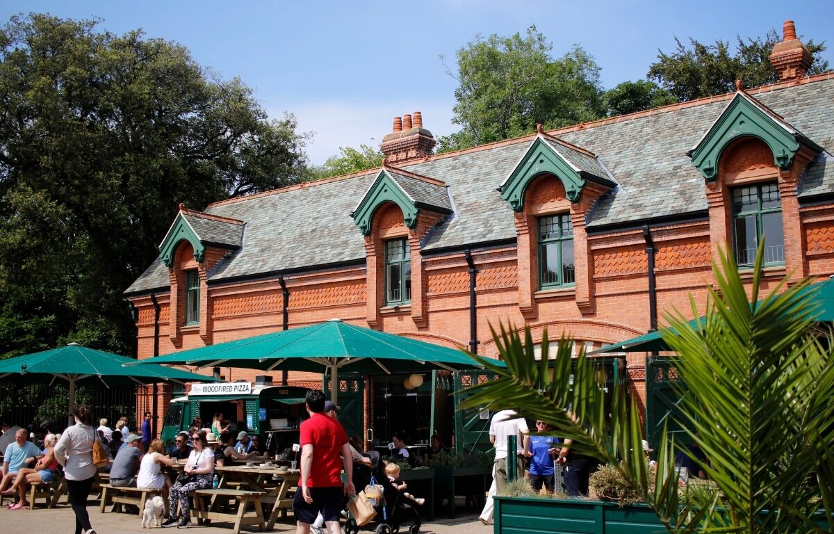 Guests enjoying outdoor dining at a charming red-brick building with green accents and lush greenery, perfect for relaxation.