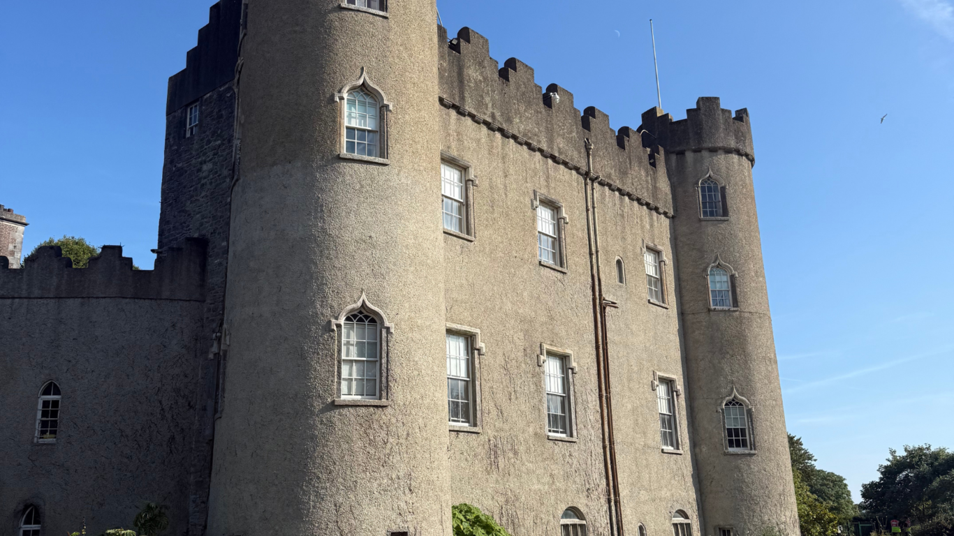 Grand historic castle hotel with elegant turrets, lush gardens, and a serene blue sky, offering a unique stay experience.