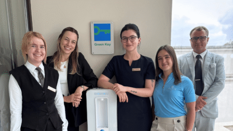 Friendly hotel staff by Green Key award sign, offering sustainable service and water station, welcoming environment.